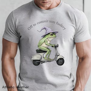 Op de afbeelding: Athletic heather grijze t-shirt met een groene kikker met een paarse tovenaars hoed, op een witte scooter. De tekst "Off to commit tom foolery" staat boven de kikker.