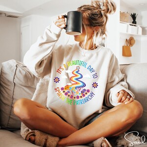 Op de afbeelding: Cr&egrave;mekleurige sweatshirt met de tekst "It's a beautiful day to smash the patriarchy" in een regenboogontwerp. De afbeelding bevat een regenboog, bloemen en wolken. Een casual, comfortabele stijl.