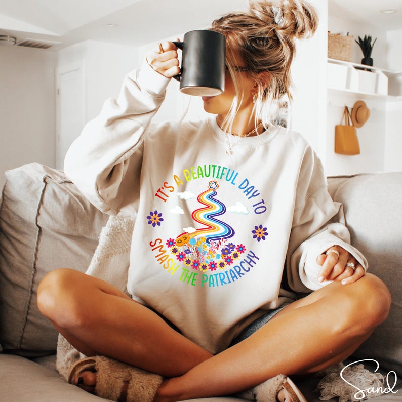 Op de afbeelding: Cr&egrave;mekleurige sweatshirt met de tekst "It's a beautiful day to smash the patriarchy" in een regenboogontwerp. De afbeelding bevat een regenboog, bloemen en wolken. Een casual, comfortabele stijl.