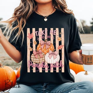 May include: A black t-shirt featuring a graphic print of three pumpkins with pink bows, surrounded by autumn leaves and vertical stripes. The text reads "TAKE ME TO THE PUMPKIN PATCH" in pink lettering. The design includes small star accents.