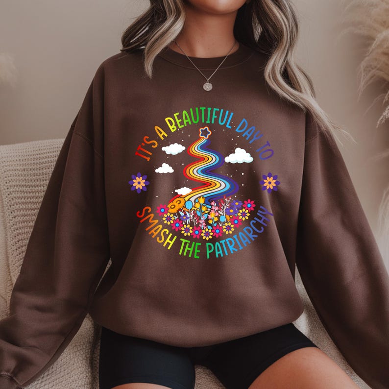 Op de afbeelding: Een bruine sweatshirt met een kleurrijke afbeelding met de tekst "It's a beautiful day to smash the patriarchy." De afbeelding toont een regenboog, bloemen, wolken en een ster.