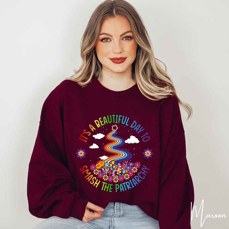 Op de afbeelding: Bordeaux sweatshirt met de tekst "It's a beautiful day to smash the patriarchy". Het ontwerp toont een regenboog, bloemen, wolken en sterren. De sweatshirt heeft een ronde hals.