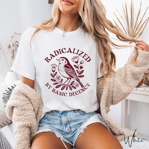 Puede incluir: Camiseta blanca con un diseño burdeos de un pájaro rodeado de flores y el texto "RADICALIZED BY BASIC DECENCY". La camiseta es de material suave, adecuada para uso diario.