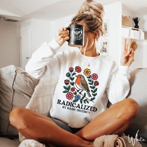 Op de afbeelding: Witte sweatshirt met een bloemenkransontwerp, een vogelillustratie en de tekst "RADICALIZED BY BASIC DECENCY". Een zwarte mok met een bijen- en maanontwerp wordt omhoog gehouden.