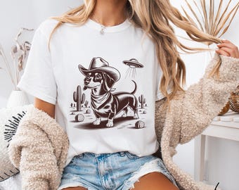 May include: White t-shirt featuring a dachshund wearing a cowboy hat and bandana, with a UFO hovering above. The graphic is in brown tones, with cacti and desert elements. The word "White" is written in script at the bottom right.