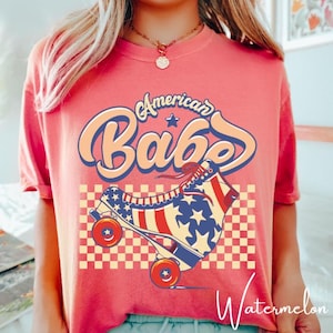 Może przedstawiać: Koralowa koszulka z motywem retro. Tekst "American Babe" jest napisany zabawną czcionką. Grafika zawiera rolkę z czerwonym, białym i niebieskim wzorem w gwiazdy i paski na szachownicy.