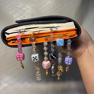 May include: Five decorative metal bookmarks attached to the pages of a closed notebook. Each bookmark features a unique arrangement of colorful beads and a single die, including pink, white, black, blue, and purple dice with various metallic charms.