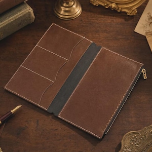 May include: A brown leather notebook cover featuring a dark gray spine, white stitching, and a gold-toned zipper. The interior includes three card slots on the left side and a full-length pocket on the right side, designed for organizing documents.
