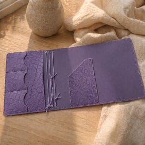 May include: Open, flat, purple leather journal with a crocodile-textured pocket and card slots. The journal has a matching elastic closure and a smooth, matte finish. The journal is open on a wooden surface.