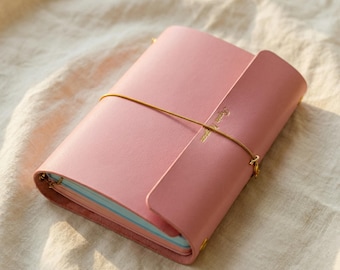 May include: A pink leather journal with a flap closure and a gold elastic band. The journal features a gold inscription on the flap and gold corner accents. The pages have a light blue hue.