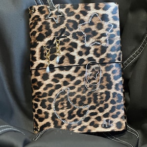 May include: A rectangular notebook with a brown and black leopard print cover. The cover is decorated with a silver star clip, a silver star-shaped paperclip, a crescent moon outline made of rhinestones, and two gold-toned dangling pearl charms.