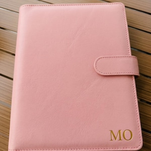 May include: A pink leather journal with a tab closure. The journal has the gold letters "MO" on the bottom right corner. The journal is closed and has a stitched border.