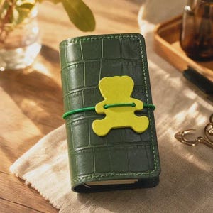 May include: A dark green leather journal with a textured, crocodile-like pattern. A yellow teddy bear-shaped embellishment is attached with a green elastic band. The journal is closed and resting on a light-colored cloth.
