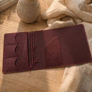 May include: Open burgundy leather journal with a crocodile-textured pocket and card slots. The journal is open, revealing a smooth interior and elastic closure cords. The journal is on a wooden surface.