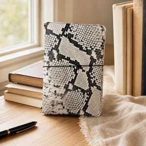 May include: A rectangular notebook cover with a snakeskin pattern in black and white. The cover is secured with a black elastic band. A black pen with gold accents rests on a wooden surface near the notebook.