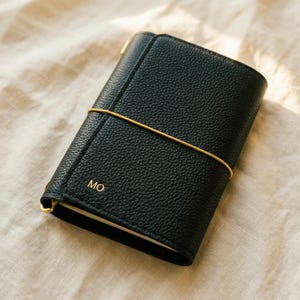 May include: A black leather journal with a textured surface, secured by a gold elastic band. The initials "MO" are embossed in gold on the front. The journal is closed and appears to be on a cream-colored fabric.