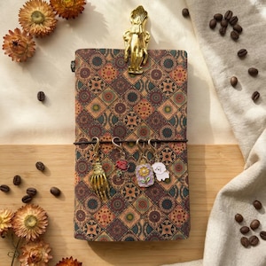 May include: A patterned journal with a gold-coloured clip and three charms. The journal has a brown, red, and green geometric design. Charms include a skeleton hand, a rose, a sunflower, and a white dog. Dried flowers and coffee beans are scattered around.