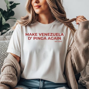 May include: Cream-colored t-shirt with the red text "MAKE VENEZUELA D' PINGA AGAIN" and "EST. 2026". The shirt is a crew neck style. The person is wearing a beige cardigan.