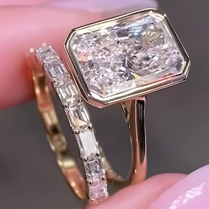 Puede incluir: Un conjunto de anillos de oro con una gran piedra central rectangular de corte radiante en un engaste de bisel, junto con una banda a juego incrustada con una hilera de diamantes más pequeños de corte baguette y acabado pulido.