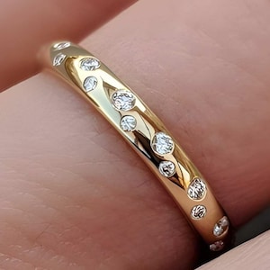 Könnte beinhalten: Ein polierter Goldring mit mehreren kleinen, runden, klaren Steinen, die in einem unregelmäßigen, verstreuten Muster bündig in die Oberfläche eingelassen sind. Das Metall hat eine glatte, reflektierende Oberfläche, die das Licht einfängt.