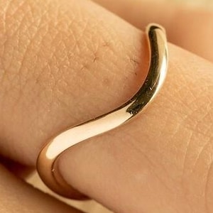 May include: A polished gold-toned ring featuring a smooth, continuous wave-like design that curves gently along the band. The metal surface is reflective and uniform in colour, showcasing a simple, curved silhouette that wraps around the finger.