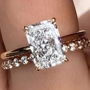 Puede incluir: Un conjunto de anillos de oro con una gran piedra central rectangular de corte radiante sujeta por cuatro garras, junto a una banda a juego adornada con una hilera de pequeñas piedras redondas de corte brillante en el lateral, creando un efecto superpuesto.