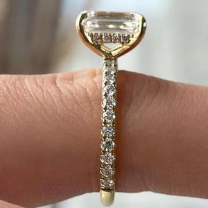 May include: A gold ring featuring a large emerald-cut centre stone held in a four-prong setting. The band is adorned with a row of small, round, clear pavé-set stones that extend along the shank, complemented by a hidden halo of small stones.