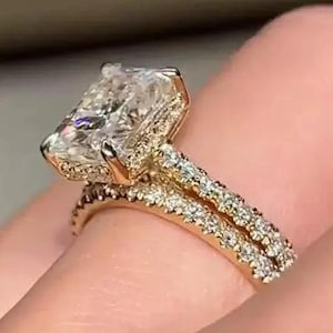 May include: A gold engagement ring with a large, rectangular-cut diamond centre stone. The band is also gold and features smaller diamonds. The ring is shown on a finger.