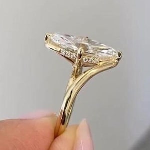 May include: A gold engagement ring featuring a large, clear, marquise-cut gemstone. The ring has a delicate band and is set with smaller accent stones. The ring is held up against a neutral background, showcasing its elegant design.