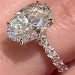 Op de afbeelding: Een ring met een grote, ovaal geslepen, heldere, gefacetteerde edelsteen die door meerdere pootjes wordt vastgehouden. De band is gemaakt van een metaalachtig materiaal en is bezet met een reeks kleinere, ronde, heldere stenen aan de zijkant.