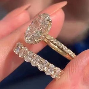 Könnte beinhalten: Ein goldener Verlobungsring mit einem großen, oval geschliffenen Diamanten und einem diamantbesetzten Band. Ein passendes Diamantband ist ebenfalls vorhanden. Die Ringe werden vor einem unscharfen Hintergrund gehalten.