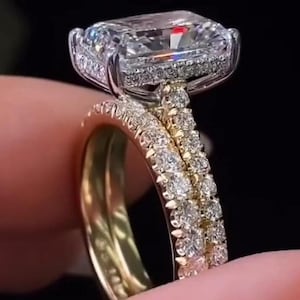 Può includere: Un anello di fidanzamento con diamante taglio radiante, una grande pietra centrale rettangolare circondata da un alone di piccoli diamanti rotondi. La fascia è in oro giallo ed è incastonata con una fila di diamanti rotondi.