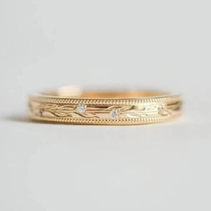 Op de afbeelding: Een gouden bandring met een gegraveerd bloemmotief en drie kleine, heldere edelstenen. De ring heeft een delicaat ontwerp met een gepolijste afwerking, geschikt voor dagelijks gebruik of speciale gelegenheden.