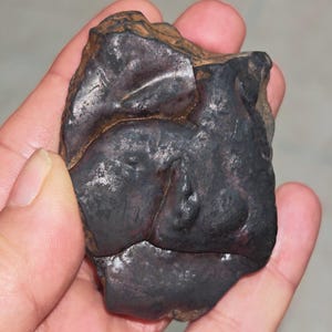 Può includere: Una roccia scura di forma irregolare con una superficie ruvida e strutturata. La roccia presenta un mix di tonalità grigio scuro e nero con alcune aree più chiare e metalliche. L'oggetto è tenuto in una mano.