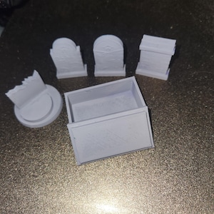 Puede incluir: Un conjunto de lápidas en miniatura y una pequeña caja, impresas en 3D en color gris claro. Las lápidas tienen diseños variados, con arcos en la parte superior y detalles grabados. La caja es rectangular con tapa. Los objetos están sobre una superficie oscura.
