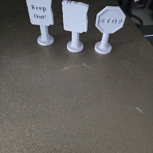 Pode incluir: Três pequenos sinais brancos com bases. O primeiro sinal diz "Keep Out!". O segundo sinal está em branco. O terceiro sinal é um sinal de stop. Todos os três sinais estão em uma superfície escura.