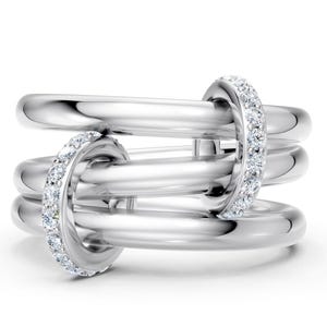 May include: A silver-colored ring with a unique design. The ring features three interconnected bands, with two bands adorned with small, sparkling stones. The ring has a polished, reflective surface, creating a modern and elegant look.