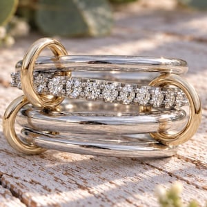 May include: A multi-band ring featuring three smooth silver-toned bands and one central band encrusted with a row of small, clear, round-cut stones. Two polished gold-toned circular links connect the bands together on the left and right sides.