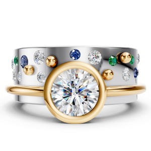 May include: A set of three rings. The primary ring features a large, round, clear gemstone set in a gold bezel. The second ring is a silver band with gold accents and is adorned with small, colorful gemstones and gold beads. The third ring is a thin gold band.
