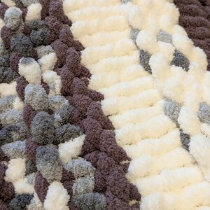 May include: Close-up of a chunky knit blanket in a mix of colors. The blanket features sections of white, gray, and dark purple yarn. The texture is soft and plush, with a thick, looped design. The overall look is cozy and inviting.