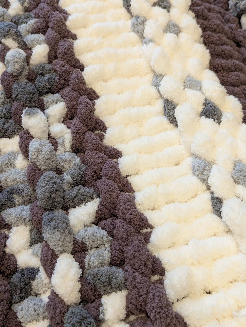 May include: Close-up of a chunky knit blanket in a mix of colors. The blanket features sections of white, gray, and dark purple yarn. The texture is soft and plush, with a thick, looped design. The overall look is cozy and inviting.