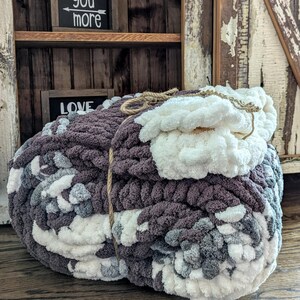 May include: Two chunky knit blankets in shades of gray, white, and purple. The blankets are tied together with a natural twine. The background includes a wooden shelf with the words "love you more" and "love" on small black signs.