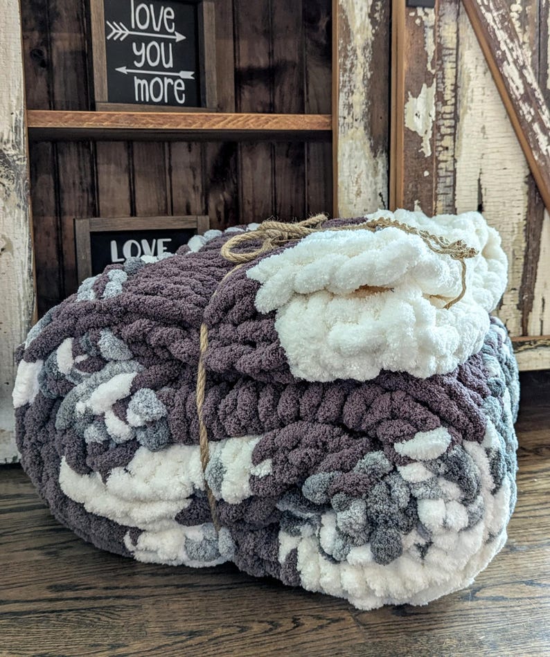May include: Two chunky knit blankets in shades of gray, white, and purple. The blankets are tied together with a natural twine. The background includes a wooden shelf with the words "love you more" and "love" on small black signs.