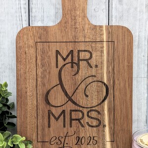 May include: Wooden cutting board with a handle, engraved with "MR. & MRS. est. 2025" in a rectangular frame. The board is a warm brown color, ideal for kitchen decor or a wedding gift.