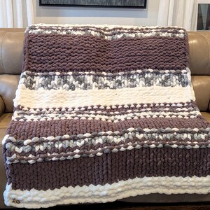 May include: A chunky knit blanket in shades of brown, white, and gray. The blanket features horizontal stripes of varying widths and textures, creating a cozy and inviting look. The soft, plush material is perfect for snuggling up on the couch.