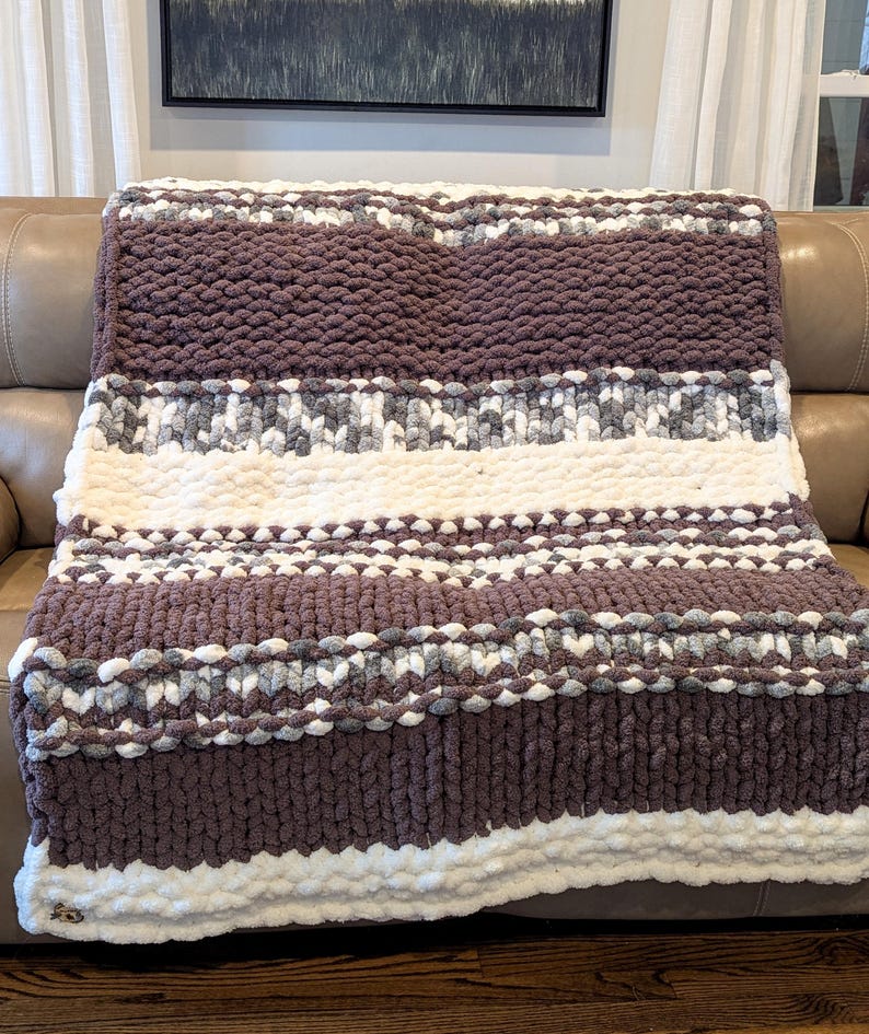 May include: A chunky knit blanket in shades of brown, white, and gray. The blanket features horizontal stripes of varying widths and textures, creating a cozy and inviting look. The soft, plush material is perfect for snuggling up on the couch.