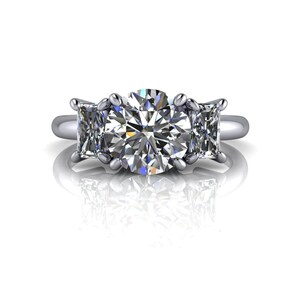 Pu&ograve; includere: Un anello di fidanzamento a tre pietre con un grande diamante rotondo centrale e due diamanti quadrati pi&ugrave; piccoli su entrambi i lati. L'anello &egrave; incastonato su una fascia in metallo bianco.