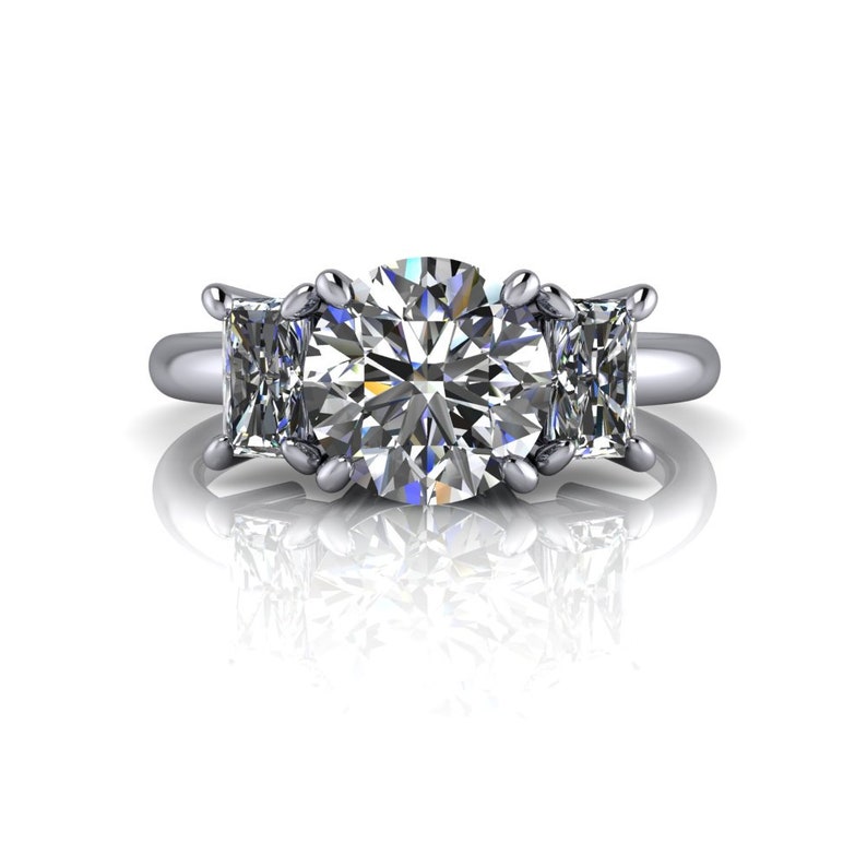 Pu&ograve; includere: Un anello di fidanzamento a tre pietre con un grande diamante rotondo centrale e due diamanti quadrati pi&ugrave; piccoli su entrambi i lati. L'anello &egrave; incastonato su una fascia in metallo bianco.