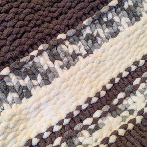 May include: A close-up of a chunky knit blanket in shades of brown, white, and gray. The blanket features diagonal stripes of varying widths, creating a textured and cozy appearance. The thick yarn gives the blanket a plush, inviting look.