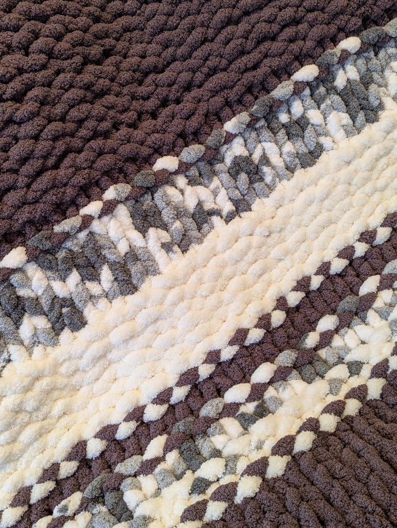 May include: A close-up of a chunky knit blanket in shades of brown, white, and gray. The blanket features diagonal stripes of varying widths, creating a textured and cozy appearance. The thick yarn gives the blanket a plush, inviting look.